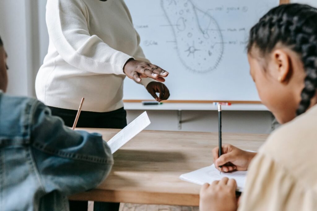 Crop faceless female teacher standing near desk while schoolchildren writing task on papers during lesson