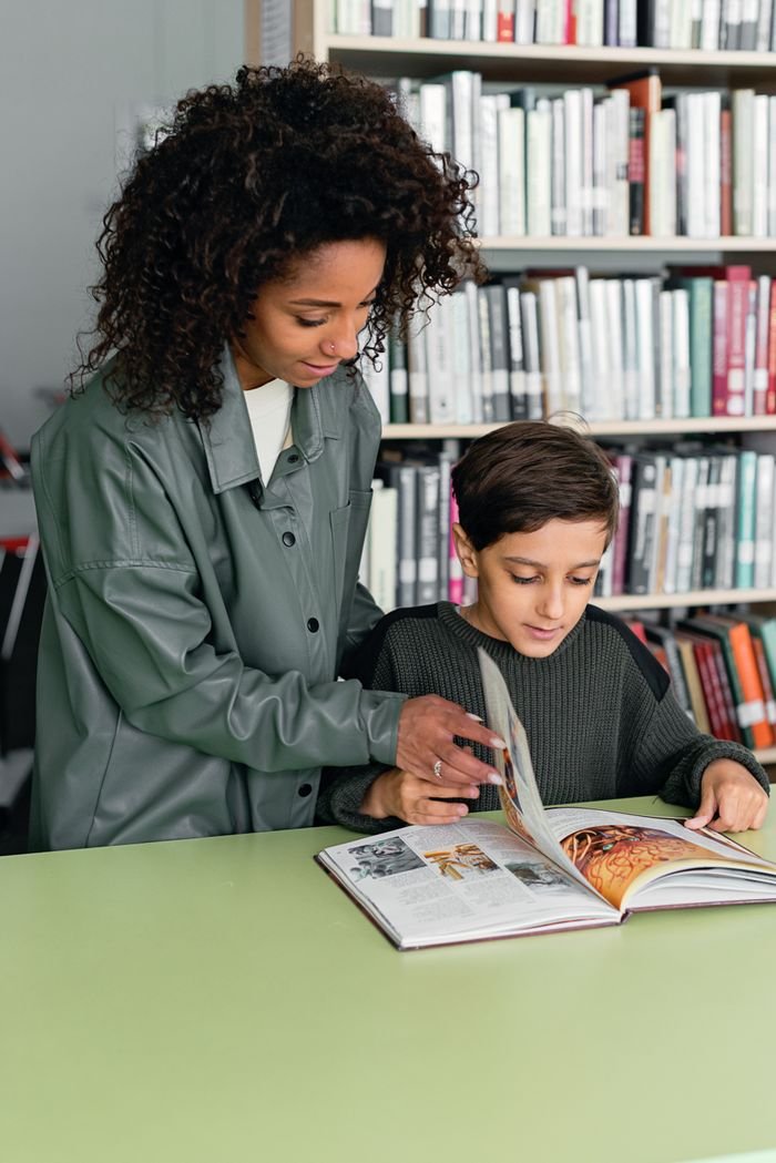 A woman assists a young boy with reading a book in a vibrant modern library setting.